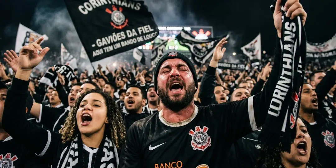 Torcedores fervorosos gritando as músicas e cantos mais famosos da torcida do Corinthians em um estádio lotado. Em destaque, um homem emocionado segura um cachecol do clube, cercado por bandeiras da Gaviões da Fiel e outros torcedores uniformizados em preto e branco.