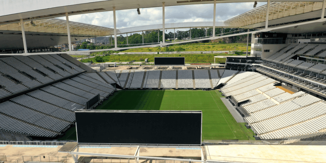 Vista panorâmica de ângulo elevado do interior da Neo Química Arena, mostrando o gramado verde impecável, as arquibancadas laterais cobertas e os grandes telões pretos desativados sob um céu parcialmente nublado.